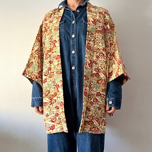 Puede incluir: Un kimono color crema con un estampado floral en rojo, verde y dorado. El kimono se lleva sobre un mono vaquero. Las mangas son anchas y la prenda está abierta por delante.