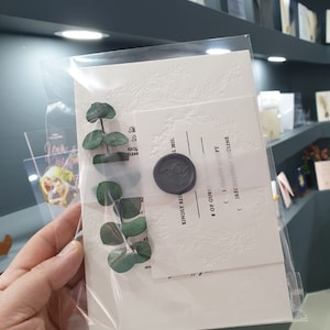 May include: A white invitation card with a wax seal and green eucalyptus leaves. The card has a decorative embossed pattern and the text "KINDLY RSVP" and "# OF GUESTS".