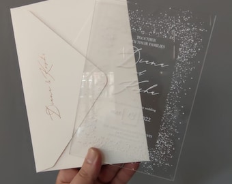 May include: A clear acrylic wedding invitation with a white glitter design. The invitation features the names "Diana & Josh" and the date "May 13, 2022".