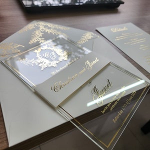May include: A sophisticated wedding invitation set. The clear acrylic invitation has gold lettering with the names "Christian and Janet" and the date "Saturday 17th of June 2023". The set includes a matching envelope and details card, all with gold accents.