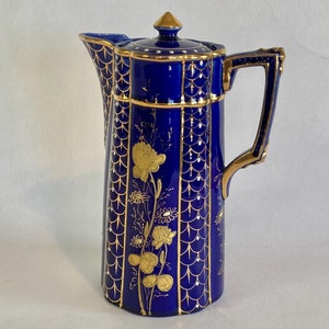 May include: A tall, cobalt blue ceramic pitcher with a lid, handle, and spout. The pitcher is decorated with gold floral and geometric patterns. The handle and rim are trimmed in gold. The pitcher is likely for serving beverages.