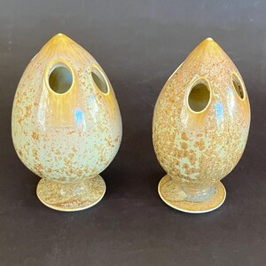 May include: Two speckled, teardrop-shaped ceramic vases with three circular openings. The vases have a light green and brown color scheme with a glossy finish. They sit on a small, round base.