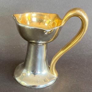May include: A small, decorative cup with a gold interior and handle. The exterior is a dark, metallic color. The cup has a flared base and a wavy rim. The handle is curved and gold. The cup is likely made of ceramic or metal.
