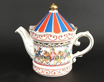 May include: A white ceramic teapot with a carousel design. The lid is striped red and blue, topped with a gold finial. The body features a carousel scene with riders on horses. The handle and spout are white with gold trim.