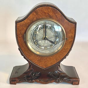 May include: A wooden mantel clock with a shield-shaped frame. The clock face is round with a gold-colored bezel and black numerals. The clock is set on a decorative base with carved details.