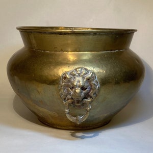 May include: A brass planter with a rounded body and a wide rim. A decorative lion's head with a ring handle is attached to the front. The planter has a tarnished finish, giving it an antique appearance.