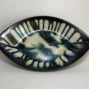May include: A ceramic leaf-shaped dish with a dark blue rim and a cream and blue interior. The dish has a glossy finish and a unique, organic design. The dish is approximately 10 inches long.