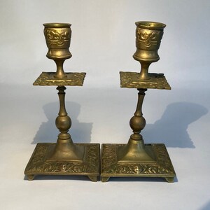 May include: Two antique brass candlesticks. Each candlestick has a square base with ornate floral detailing, a slender stem, a square platform, and a cup-shaped top. The candlesticks are a warm gold color and are displayed against a white background.
