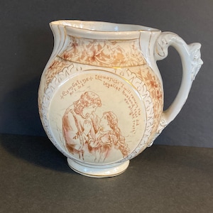 May include: Cream-colored antique pitcher with a detailed handle and gold trim. The pitcher features a sepia-toned illustration of a couple and text from Shakespeare's Cymbeline. Floral patterns and additional gold accents adorn the pitcher's surface.