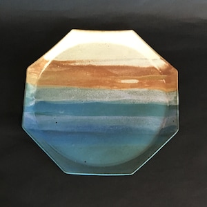 May include: An octagonal ceramic plate with a painted landscape design. The plate features horizontal bands of color, including beige, brown, teal, and blue. The plate has a clear, glossy finish.