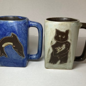 May include: Two ceramic mugs. One is blue with a brown dolphin design. The other is off-white with a brown cat holding a bird. Both mugs have square handles and brown interiors.