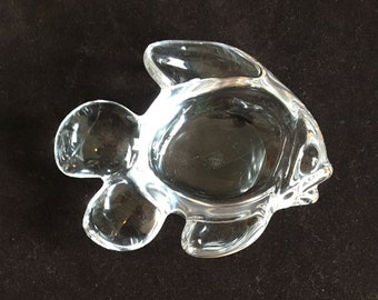May include: A clear glass fish-shaped dish with a central bowl and fin-like extensions. The dish is transparent, allowing light to pass through, and sits on a dark surface. The fish design adds a decorative element.