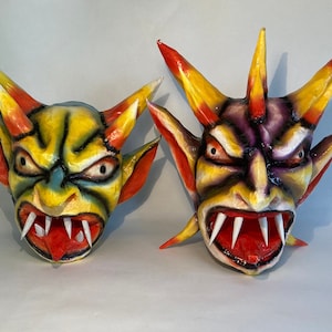 May include: Two handmade, colorful demon masks. One is yellow, green, and orange, the other is purple, red, and yellow. Both have sharp teeth, red tongues, and menacing expressions. The masks are likely for Halloween or costume use.