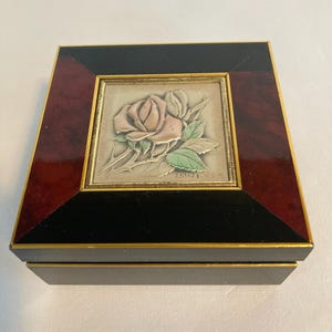 May include: A square jewelry box with a rose design. The box has a black base and a frame with a dark red wood grain pattern. The center features a framed image of a pink rose with green leaves.