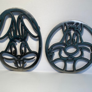 May include: Two dark teal metal trivets featuring cartoon character faces. One is oval-shaped with Bugs Bunny's face, and the other is round with a cartoon cat's face. Both have openwork designs.