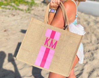 May include: A woman wearing a straw hat and sunglasses is holding a large, natural-fiber tote bag with pink stripes and the initials "KM" printed on it.