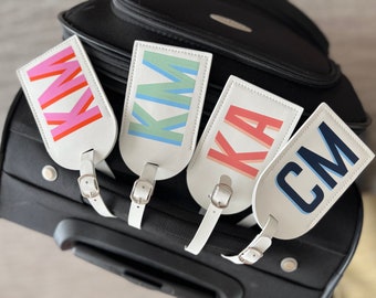 May include: Four white luggage tags with colorful letters spelling out the names KIM, KA, and CM. The tags are attached to a black suitcase.