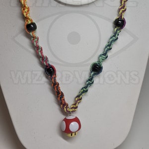 May include: A colorful necklace with a red and white mushroom pendant. The necklace features a braided cord in rainbow hues, interspersed with black beads. The pendant has a gold-colored base.