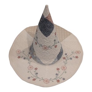 May include: A conical hat constructed from quilted fabric featuring a patchwork design in shades of cream, navy blue, and dusty rose. The wide brim and pointed crown are decorated with embroidered floral vines in red, blue, and green thread.