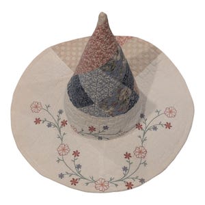 May include: A pointed hat constructed from quilted fabric featuring a patchwork cone in shades of dusty pink, blue, and cream. The wide, cream-colored brim is decorated with embroidered floral vines in red and blue thread, showcasing detailed stitching.
