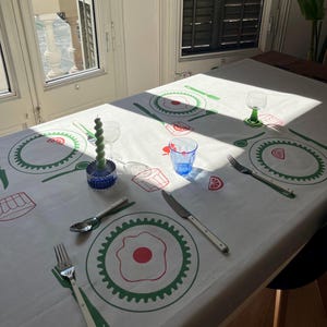 May include: A white tablecloth with a green and red design featuring stylized plates, glasses, and cutlery. The design includes a fried egg, a lemon slice, and a glass of juice.