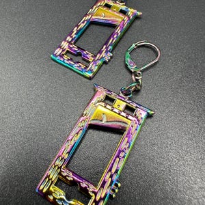May include: A pair of iridescent guillotine-shaped earrings. The earrings feature a rectangular frame with a blade and are suspended from silver-colored ear wires. The metal has a rainbow finish, with colors shifting from gold to blue and purple.