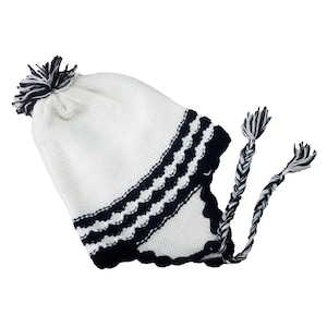 Könnte beinhalten: Eine weiße Strickmütze mit einem schwarz-weißen Bommel und Ohrenklappen. Die Mütze hat eine schwarze, gewellte Bordüre und schwarz-weiße geflochtene Bänder. Die Mütze soll den Träger bei kaltem Wetter warm halten.