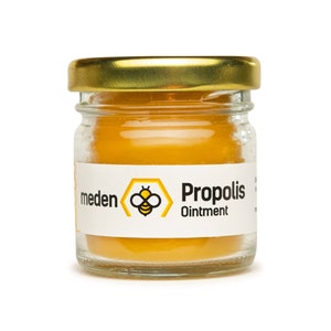 May include: A small glass jar with a gold lid containing yellow ointment. The jar has a white label with the words "meden" and "Propolis Ointment" and a bee logo. This is likely a skincare product.