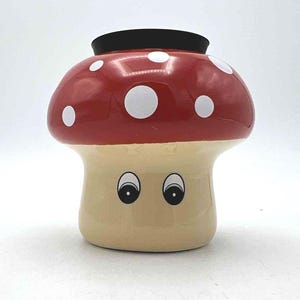 May include: A whimsical mushroom-shaped ceramic container. The mushroom cap is red with white polka dots, and the stem is beige. It has two cartoon eyes. The top has a black rim.