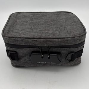 May include: A gray, rectangular storage case with a combination lock. The case has a textured fabric exterior, a black zipper, and black handles on each side. The combination lock is black with white numbers.