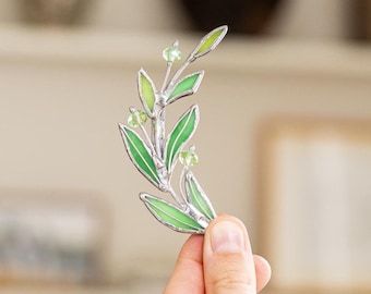 May include: A stained glass suncatcher shaped like a curved leafy branch with light green glass leaves and small clear glass beads, held upright. The piece features silver-toned metal soldering outlining each individual leaf and stem segment.