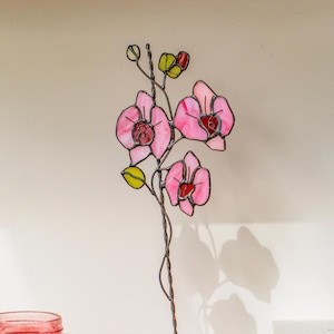 Può includere: Una scultura di orchidea in vetro colorato con petali rosa e dettagli verdi. Il fiore è montato su una base di legno, creando un elemento decorativo. La scultura è esposta su una mensola di legno con altri oggetti decorativi.