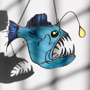 Puede incluir: Una suspensión de vidrio coloreado que representa un pez pescador azul con un señuelo amarillo brillante. El pez tiene dientes afilados y una boca grande.