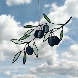 Peut inclure: Une branche d'olivier en verre coloré avec des feuilles vertes et des olives noires suspendue à une ventouse contre un ciel bleu avec des nuages blancs.