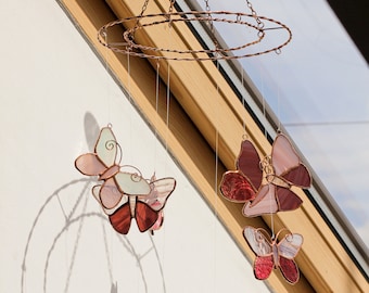 Puede incluir: Un móvil de mariposas de vidrio coloreado con cinco mariposas en tonos de rosa, rojo y blanco que cuelgan de un marco de alambre de cobre.