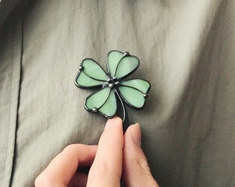 May include: A four-leaf clover brooch crafted from light green stained glass with dark metal soldering lines. The clover features four rounded petals and a small stem, pinned to an olive green button-down shirt worn over a white t-shirt.