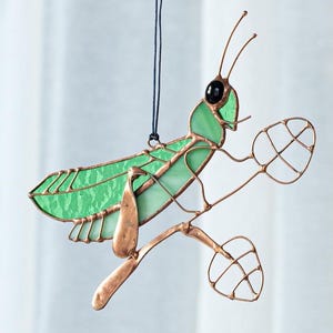 May include: A stained glass praying mantis suncatcher. The insect features a green body, copper-colored legs and antennae, and a black eye. It is suspended by a black string, and is perched on a copper-colored branch with two wire-framed leaves.