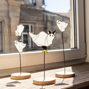 May include: A set of whimsical Halloween decorations featuring white ghost and cat figures. The figures are mounted on thin black wire stems and set on round wooden bases. One cat figure is perched on a yellow crescent moon.
