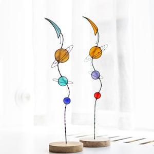 Pode incluir: Duas esculturas verticais de vidro colorido com arranjos planetários em hastes metálicas finas. Cada peça tem uma base circular de madeira e exibe esferas de vidro coloridas com anéis de arame, encimadas por uma forma de meia-lua.