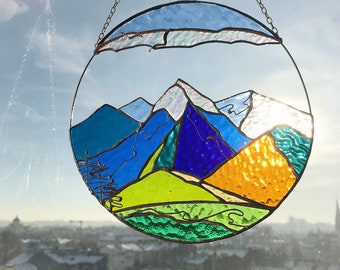 May include: A round stained glass window hanging with a chain. The window depicts a mountain range with a blue sky and green hills. The mountains are made of blue, green, yellow, and white glass.