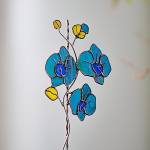 May include: A stained glass orchid sculpture with blue and yellow flower petals. The petals have a gradient of colors, from light to dark blue. The flower's center is a darker blue. The sculpture is on a twisted metal stem.