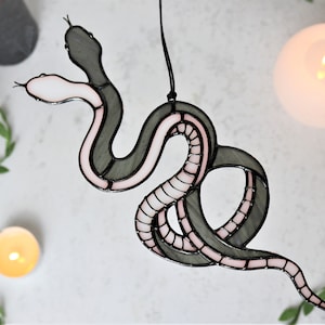 Puede incluir: Una serpiente de vidrio coloreado con un diseño rosa y gris. La serpiente está enroscada en un círculo y tiene una cola larga.