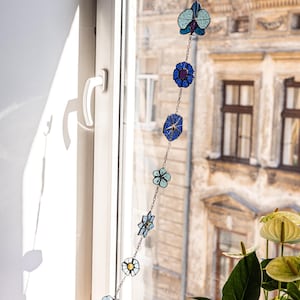 Op de afbeelding: Een decoratieve bloemenslinger van glas-in-lood in blauw, groen en geel hangt voor een raam. De slinger heeft verschillende bloemvormen die aan elkaar zijn geregen met een delicate ketting. Op de achtergrond is een gebouw te zien.