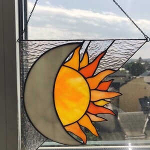 May include: A stained glass sun and moon window hanging. The sun is yellow and orange with a red outline. The moon is gray. The window is hanging from a chain.
