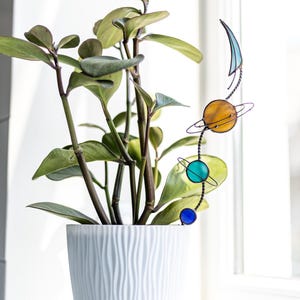 May include: A stained glass plant stake featuring a vertical arrangement of a crescent moon, a large amber planet with rings, a medium teal planet with rings, and a small blue sphere, all connected by a dark metal wire stem inserted into a white pot.