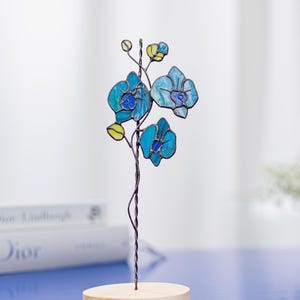May include: A stained glass orchid sculpture with blue and yellow petals, mounted on a wooden base. The flower has a twisted wire stem. The sculpture is displayed on a blue surface.