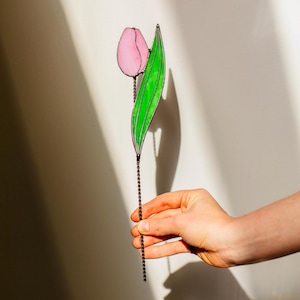May include: A stained glass tulip with a pink flower and a green leaf. The tulip is held by a dark metal chain. The artwork is set against a white background.