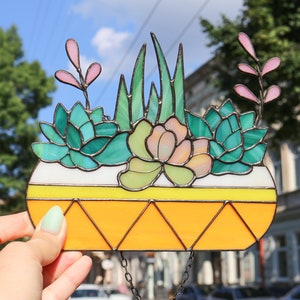 May include: A stained glass window hanging featuring a planter with succulents and a green leafy plant. The planter is orange with a white rim and yellow stripe. The background is a blurred image of a city street.