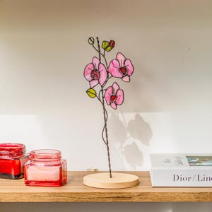 May include: A handcrafted stained glass orchid sculpture with pink petals and green accents. The flower is mounted on a thin wire stem and a round wooden base. The sculpture is displayed on a wooden shelf with other decorative items.