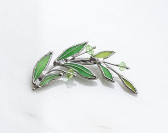 May include: A silver-toned brooch shaped like a leafy branch with vibrant green enamel filling the leaf segments. The design includes small, faceted light green beads attached to the ends of thin metal stems, creating a botanical appearance.